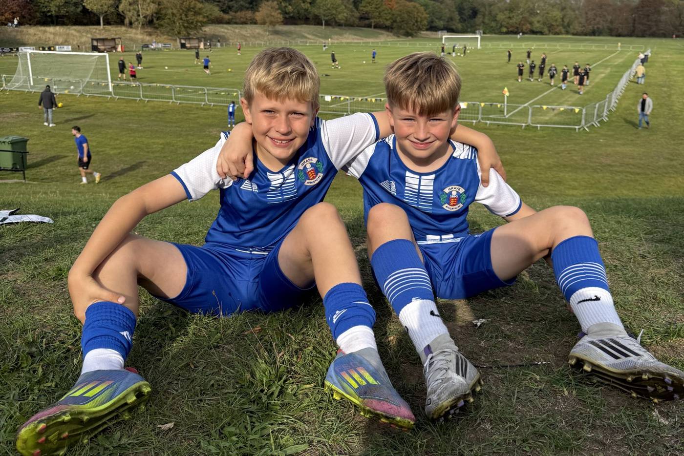 Two smiling Grays Athletic youth players sitting on the grass at Treetops, with matches being played behind them